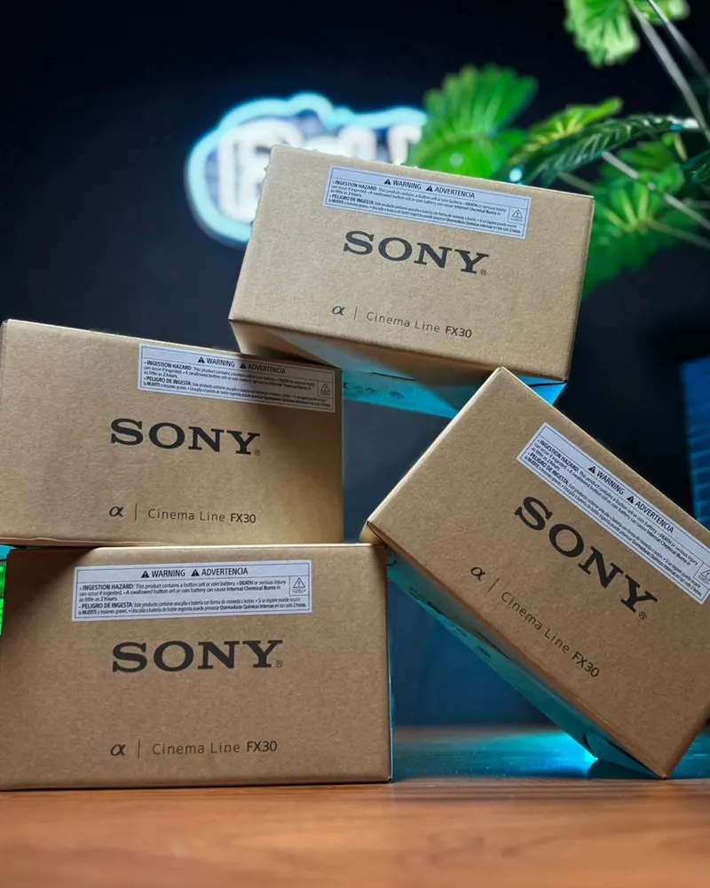 Quatro caixas lacradas da câmera Sony FX30 empilhadas numa mesa de madeira, com o letreiro neon da BeHe ao fundo
