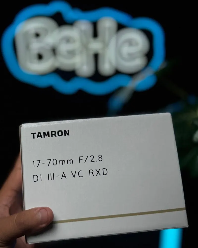Caixa da lente Tamron 17-70mm F/2.8 Di III-A VC RXD segurada na mão, em frente ao letreiro neon da BeHe